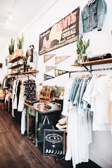 A vintage-style store interior featuring clothing racks with denim and light-colored garments. Shelves display potted plants, leather bags, and various decorative items. A large vintage Levi's sign and pennants are mounted on the wall, along with other unique decor like an old typewriter and a decorative chart with moon phases.