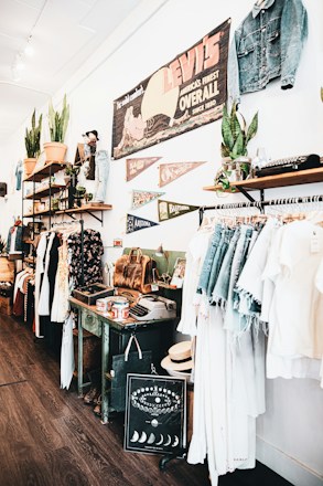 A vintage-style store interior featuring clothing racks with denim and light-colored garments. Shelves display potted plants, leather bags, and various decorative items. A large vintage Levi's sign and pennants are mounted on the wall, along with other unique decor like an old typewriter and a decorative chart with moon phases.