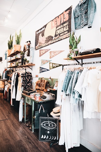 A vintage-style store interior featuring clothing racks with denim and light-colored garments. Shelves display potted plants, leather bags, and various decorative items. A large vintage Levi's sign and pennants are mounted on the wall, along with other unique decor like an old typewriter and a decorative chart with moon phases.