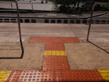 A tactile paving area on a sidewalk featuring textured surfaces in red and yellow for visually impaired pedestrians, bordered by metal railings and a street in the background.