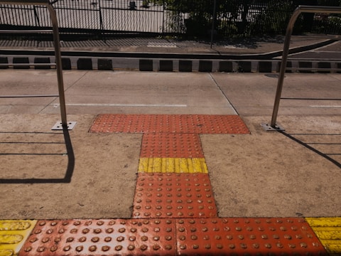 A tactile paving area on a sidewalk featuring textured surfaces in red and yellow for visually impaired pedestrians, bordered by metal railings and a street in the background.