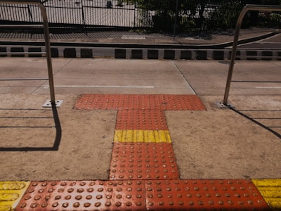 A tactile paving area on a sidewalk featuring textured surfaces in red and yellow for visually impaired pedestrians, bordered by metal railings and a street in the background.