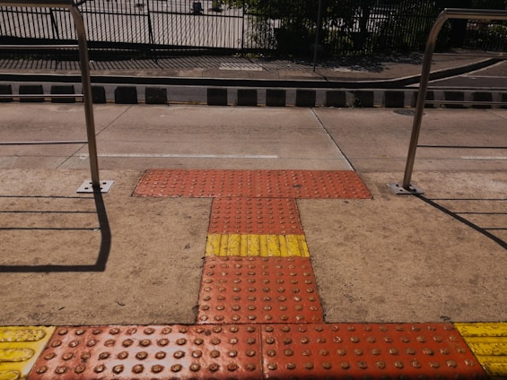 A tactile paving area on a sidewalk featuring textured surfaces in red and yellow for visually impaired pedestrians, bordered by metal railings and a street in the background.