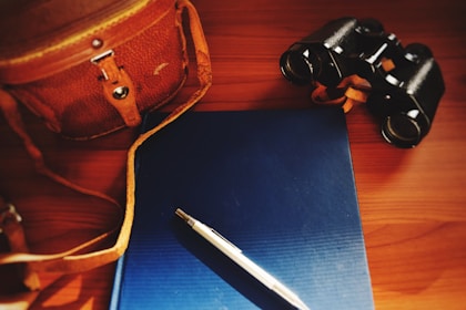 A leather bag with a strap, a blue hardcover book, a silver pen, and a pair of binoculars are arranged on a wooden surface. The lighting is warm and slightly dim, highlighting the objects with a soft glow.