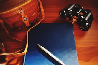 A leather bag with a strap, a blue hardcover book, a silver pen, and a pair of binoculars are arranged on a wooden surface. The lighting is warm and slightly dim, highlighting the objects with a soft glow.