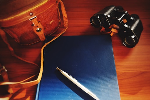 A leather bag with a strap, a blue hardcover book, a silver pen, and a pair of binoculars are arranged on a wooden surface. The lighting is warm and slightly dim, highlighting the objects with a soft glow.