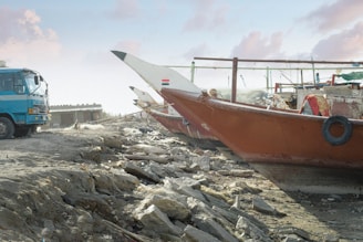 A large blue truck is parked on a rocky, uneven surface next to traditional wooden boats. The boats, with painted hulls and various details, are positioned on the dry land. The sky is partly cloudy, and there is a building structure visible in the background.