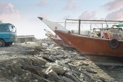 A large blue truck is parked on a rocky, uneven surface next to traditional wooden boats. The boats, with painted hulls and various details, are positioned on the dry land. The sky is partly cloudy, and there is a building structure visible in the background.