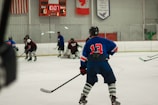 Players wearing full hockey gear are engaged in a game on an ice rink. A player in a blue jersey with the number 13 is prominently positioned in the foreground, observing the action. The scoreboard in the background shows a score of 0 for the home team and 5 for the guests with 10:07 remaining in the third period. Flags and banners hang above the rink.