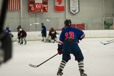 Players wearing full hockey gear are engaged in a game on an ice rink. A player in a blue jersey with the number 13 is prominently positioned in the foreground, observing the action. The scoreboard in the background shows a score of 0 for the home team and 5 for the guests with 10:07 remaining in the third period. Flags and banners hang above the rink.