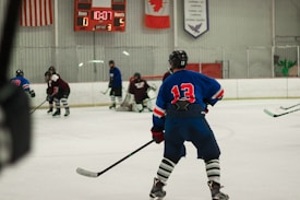 Players wearing full hockey gear are engaged in a game on an ice rink. A player in a blue jersey with the number 13 is prominently positioned in the foreground, observing the action. The scoreboard in the background shows a score of 0 for the home team and 5 for the guests with 10:07 remaining in the third period. Flags and banners hang above the rink.