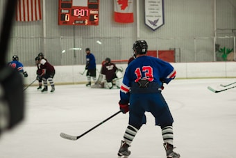 Players wearing full hockey gear are engaged in a game on an ice rink. A player in a blue jersey with the number 13 is prominently positioned in the foreground, observing the action. The scoreboard in the background shows a score of 0 for the home team and 5 for the guests with 10:07 remaining in the third period. Flags and banners hang above the rink.