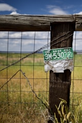 A weathered wooden fence post supports a metal grid fence with a vintage green and white license plate attached. The landscape consists of dry grass and a vast blue sky with scattered clouds.