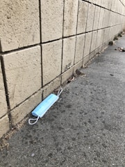 A disposable blue face mask lies on a concrete sidewalk next to a textured brick wall. The area around the mask is sparse, with some dry leaves and dirt scattered on the ground.