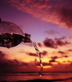 A plastic bottle tilted to pour water, positioned against a vibrant sunset sky. The water streams elegantly from the bottle, with the warm hues of orange, purple, and pink in the background creating a striking contrast.