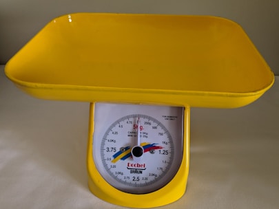 A yellow mechanical kitchen scale with a tray on top. The scale is designed to measure weights up to 5 kilograms, with measurements marked on a circular dial.