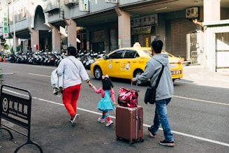 A family of three, including a child in a bright blue dress and pink accessories, walks along a city street. The adults are carrying luggage and holding hands with the child. There is a row of parked motorcycles in the background, and a yellow taxi drives by.