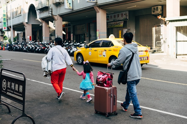 A family of three, including a child in a bright blue dress and pink accessories, walks along a city street. The adults are carrying luggage and holding hands with the child. There is a row of parked motorcycles in the background, and a yellow taxi drives by.