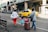 A family of three, including a child in a bright blue dress and pink accessories, walks along a city street. The adults are carrying luggage and holding hands with the child. There is a row of parked motorcycles in the background, and a yellow taxi drives by.