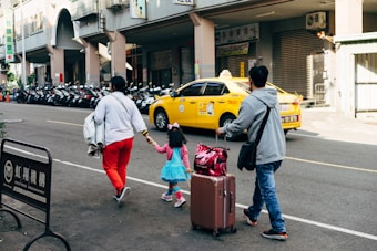 A family of three, including a child in a bright blue dress and pink accessories, walks along a city street. The adults are carrying luggage and holding hands with the child. There is a row of parked motorcycles in the background, and a yellow taxi drives by.