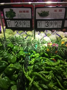 A display of various green peppers on a supermarket shelf, with price tags indicating the cost per 500g. The peppers are neatly arranged alongside other produce like cabbages. There are also some decorative flowers in the background.
