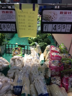 This image shows a section of a supermarket vegetable display. Various types of mushrooms, both packaged and unpackaged, are arranged on shelves with green base mats. Two prominent signs indicate prices in Chinese Yuan for different types of mushrooms; one sign lists prices for multiple types, while the other highlights the price for needle mushrooms at 4.59 Yuan per 500 grams. The display includes other vegetables like broccoli in the background.