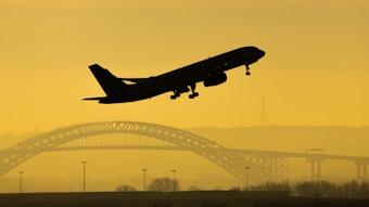 A passenger airplane is silhouetted against a yellow sky, capturing the moment it takes off. In the background, an iconic arched bridge spans across the scene, with faint outlines of hills and a few vertical structures. The image conveys a sense of travel or transition.