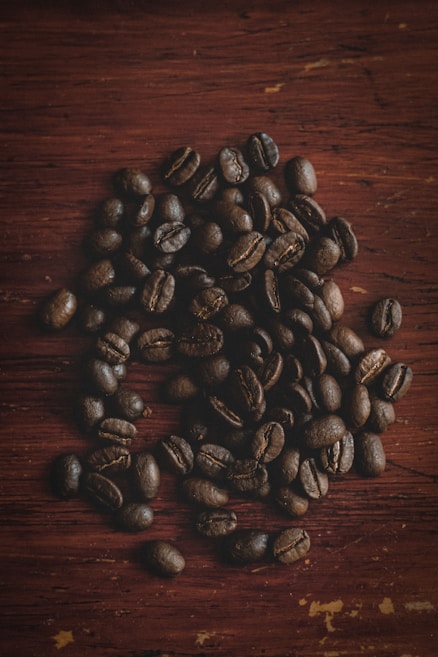 A pile of dark roasted coffee beans scattered on a rich, brown wooden surface. The texture of the beans and the wood grain are clearly visible, highlighting the rustic and earthy feel of the composition.