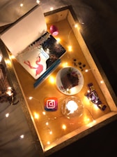 A cozy setup featuring a wooden tray adorned with string lights. Inside the tray are a notebook with a flamingo and fireworks design titled 'January', a pink oval-shaped lip balm, a plate with a piece of chocolate cake, chocolate bars, an Instagram coaster, and small coin-like objects possibly chocolates. A lit candle adds warmth to the scene.