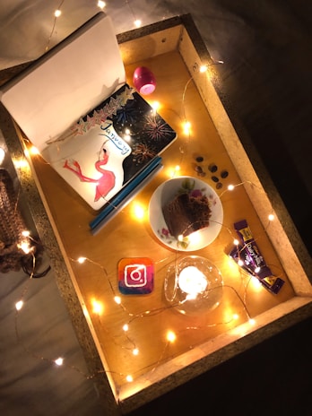 A cozy setup featuring a wooden tray adorned with string lights. Inside the tray are a notebook with a flamingo and fireworks design titled 'January', a pink oval-shaped lip balm, a plate with a piece of chocolate cake, chocolate bars, an Instagram coaster, and small coin-like objects possibly chocolates. A lit candle adds warmth to the scene.