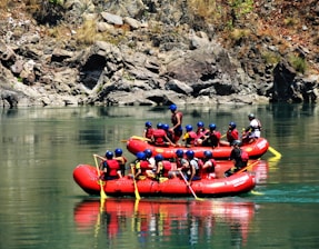 A group of people wearing helmets and life jackets engage in rafting on a calm river, navigating through rocky terrain. The scene is set against a backdrop of rugged cliffs and natural greenery.