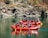 A group of people wearing helmets and life jackets engage in rafting on a calm river, navigating through rocky terrain. The scene is set against a backdrop of rugged cliffs and natural greenery.