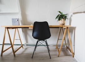 A minimalist workspace featuring a wooden desk supported by trestle legs and a sleek black chair positioned centrally. On the desk, there is a potted plant with green leaves in a white pot with brown accents, placed to the right, and a closed book with a textured gray cover near the left end. The room has light-colored wooden flooring and white walls, contributing to the minimal aesthetic.