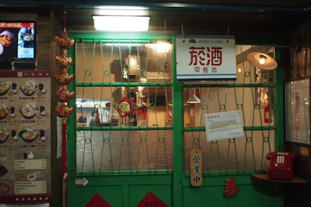 A storefront with a green metal gate and a sign with Chinese characters is visible. The interior is softly lit, showcasing various decorative items and a menu on the left side. Hanging lanterns and red ornaments add to the decor. A vintage-style red telephone is mounted on the wall next to the door.