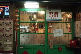 A storefront with a green metal gate and a sign with Chinese characters is visible. The interior is softly lit, showcasing various decorative items and a menu on the left side. Hanging lanterns and red ornaments add to the decor. A vintage-style red telephone is mounted on the wall next to the door.