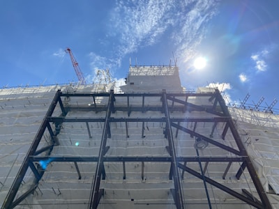 A construction site with a building covered in white protective sheeting and exposed metal framework. A red crane is visible in the background, silhouetted against a bright blue sky with the sun casting a glare.