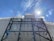 A construction site with a building covered in white protective sheeting and exposed metal framework. A red crane is visible in the background, silhouetted against a bright blue sky with the sun casting a glare.
