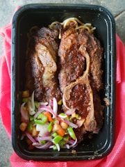 A black tray contains two pieces of cooked meat with visible brown seasoning and grill marks, accompanied by a fresh salad of diced green peppers, tomatoes, and sliced red onions. Some noodles are also visible beneath the meat. The tray rests on a red cloth.