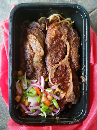 A black tray contains two pieces of cooked meat with visible brown seasoning and grill marks, accompanied by a fresh salad of diced green peppers, tomatoes, and sliced red onions. Some noodles are also visible beneath the meat. The tray rests on a red cloth.
