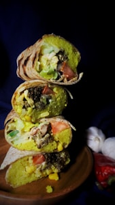 Three cross-sections of stacked burritos on a wooden surface, revealing ingredients such as lettuce, corn, tomato, rice, and some type of meat filling. In the background, there are garlic cloves and a chili pepper partially visible.