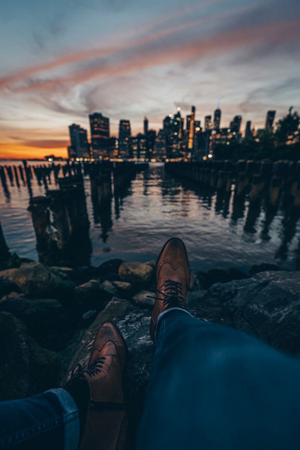 person in blue denim jeans and brown leather shoes sitting on rock near body of water