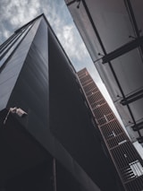 Two tall, modern buildings seen from an upward angle against a cloudy sky, with one building featuring a dark facade and the other having a metallic grid structure. A security camera is visible on the corner of the dark building.
