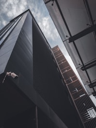 Two tall, modern buildings seen from an upward angle against a cloudy sky, with one building featuring a dark facade and the other having a metallic grid structure. A security camera is visible on the corner of the dark building.