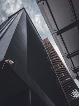 Two tall, modern buildings seen from an upward angle against a cloudy sky, with one building featuring a dark facade and the other having a metallic grid structure. A security camera is visible on the corner of the dark building.