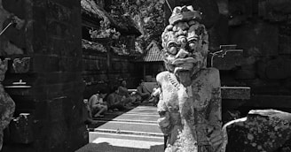 A stone statue with intricate carvings stands prominently in an outdoor setting, depicting a fierce mythological figure with exaggerated facial features and a protruding tongue. Nearby, several people are seated on steps, blending into the shadowy background under the eaves of traditional architecture. The environment suggests a historical or cultural site with rustic stone structures and a thatched roof.