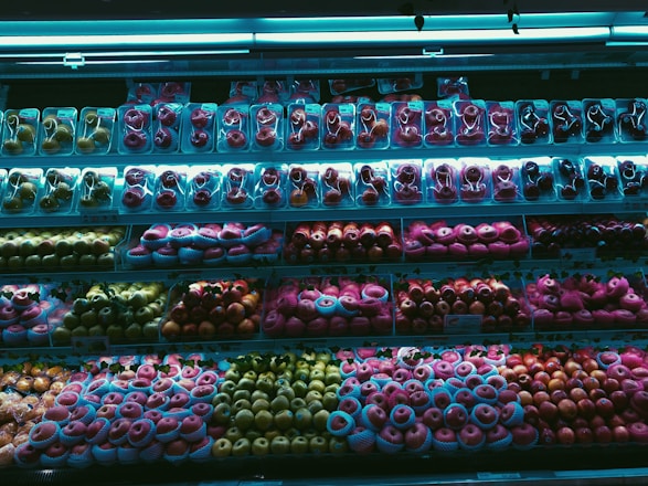 A supermarket fruit display showcases a wide variety of packaged fruits, including different types of apples and possibly other round fruits, arranged neatly across multiple shelves. The lighting casts a blue hue over the scene, highlighting the vibrant colors of the fruits.