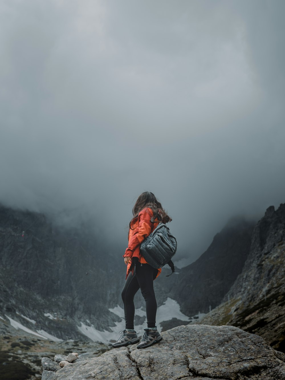 Frau in schwarzer Jacke und orangefarbenem Rucksack auf Felsformation stehend