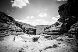 A person runs across a rugged, rocky terrain in a desert canyon under a partly cloudy sky. The cliffs and rock formations create a dramatic landscape with intricate textures and shadows.