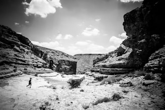 A person runs across a rugged, rocky terrain in a desert canyon under a partly cloudy sky. The cliffs and rock formations create a dramatic landscape with intricate textures and shadows.