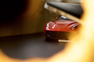A red sports car partially visible through a blurred circular opening, possibly taken from a unique angle, enhancing the depth and focus on the car's front grille and sleek design.